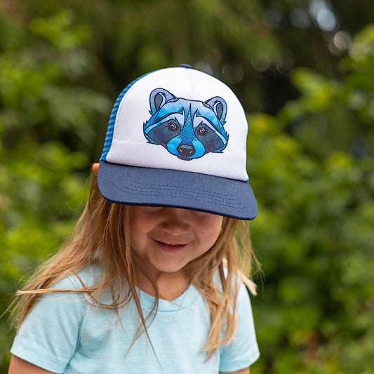 Toddler wearing a trucker hat with a navy brim, blue back and blue raccoon face design on the white front panel.