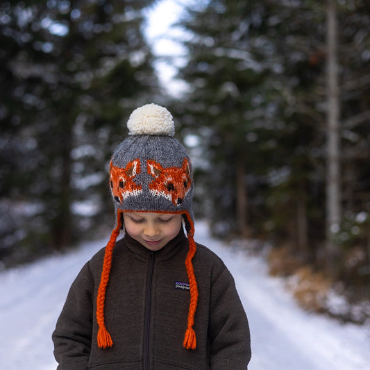 Boy wearing Ambler Creatures handknit kids' toque in heather grey with knitted fox faces, coloured tassels and pom.