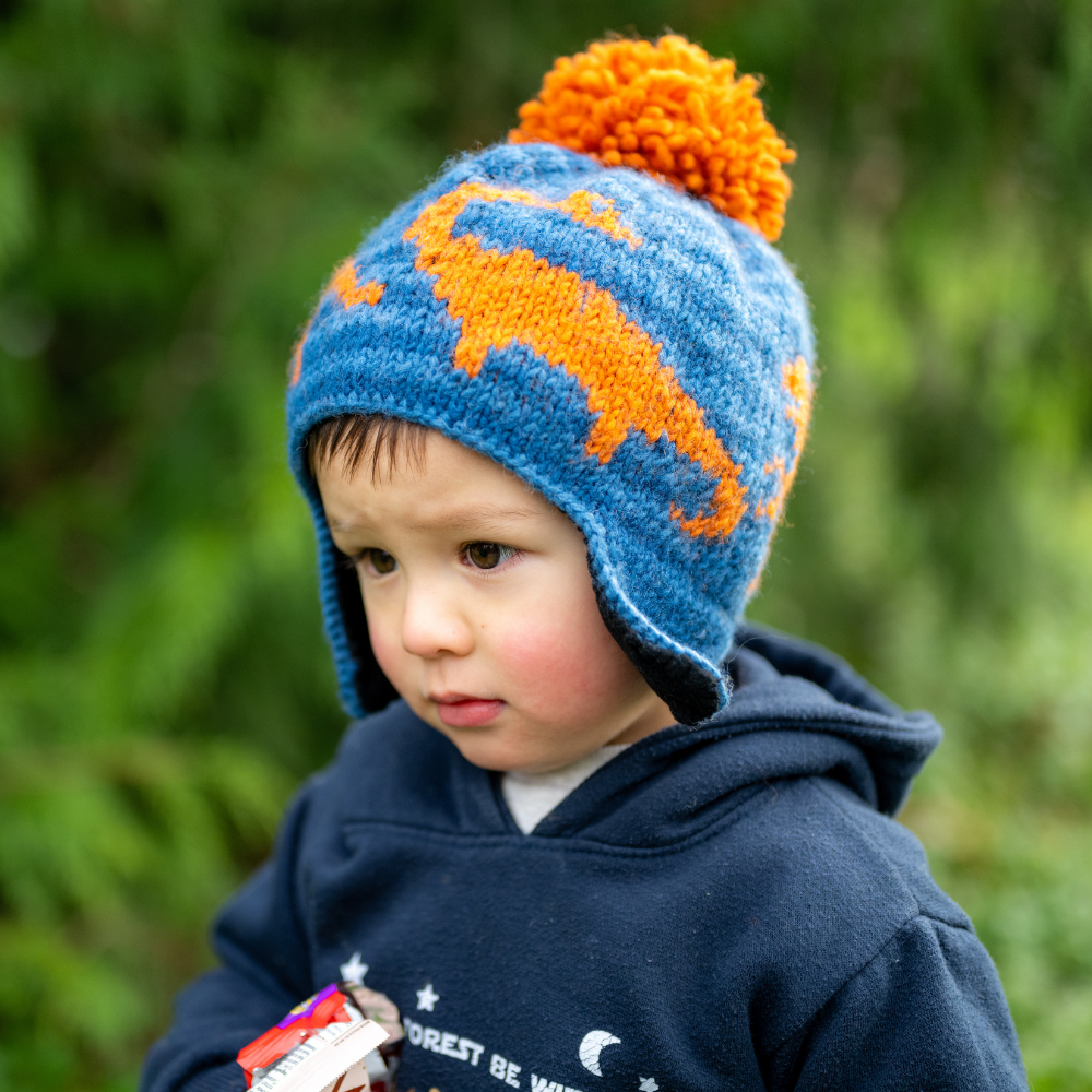 Boy wearing Ambler Dino handknit kids' toque in blue with embroidered dinosaur pattern and contrast colour pom.