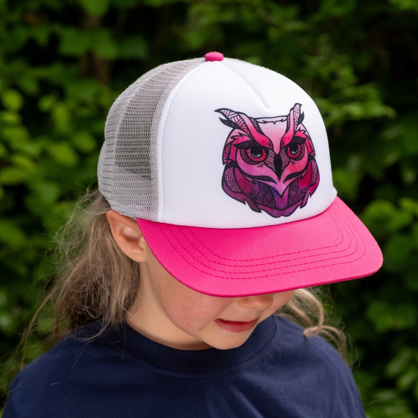 Toddler girl  wearing a trucker hat with a pink brim, white back and pink owl face design on the white front panel. 