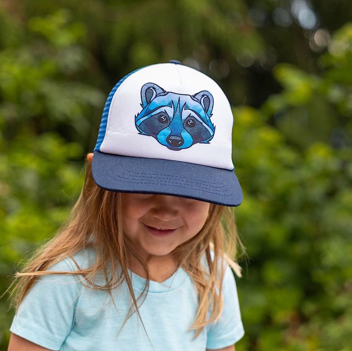 Toddler wearing a trucker hat with a navy brim, blue back and blue raccoon face design on the white front panel. 