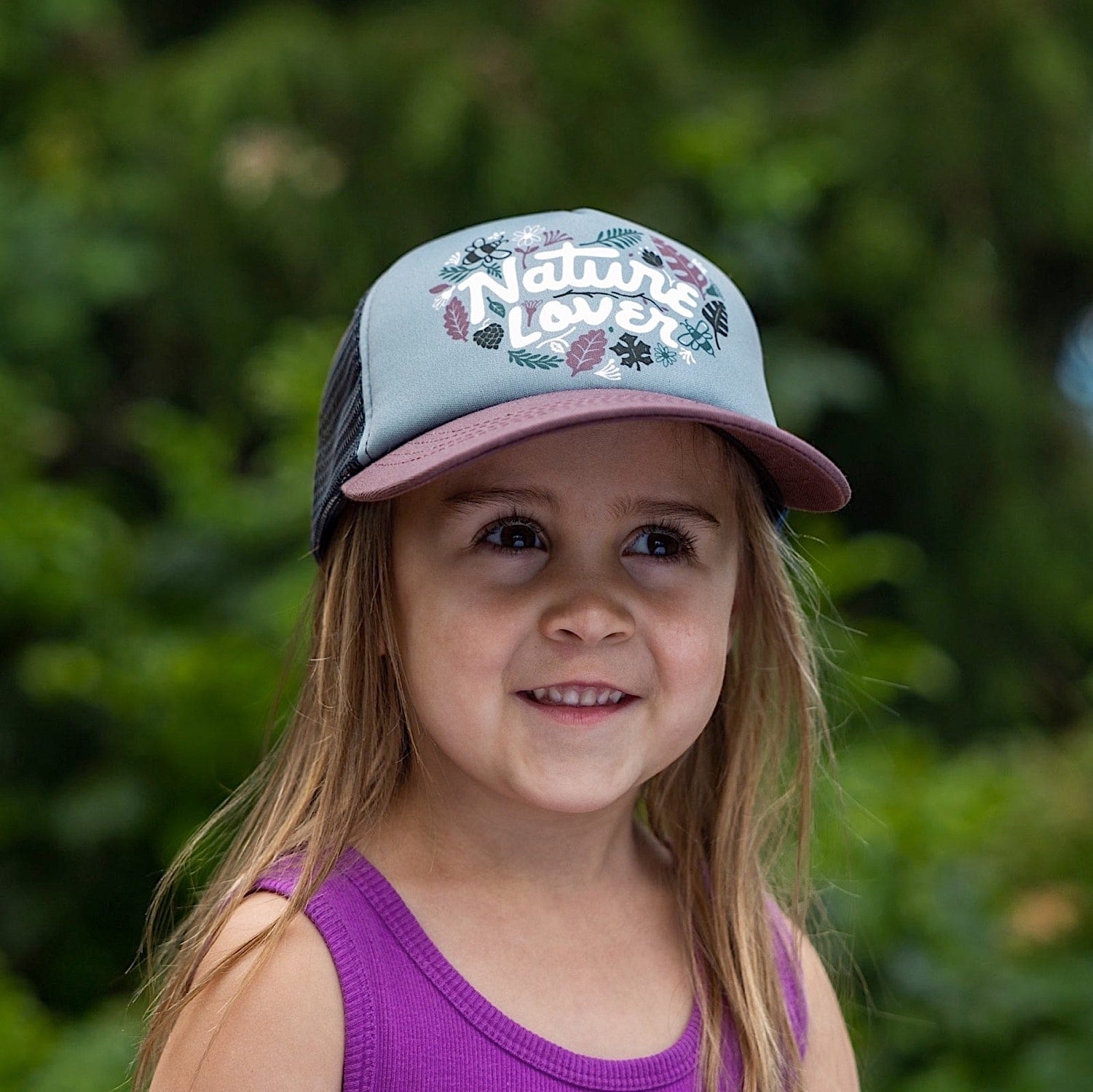 Girl wearing a kids' trucker hat with purple brim, black mesh back and a 'Nature Lover' text and leaf graphic on the front panel. 