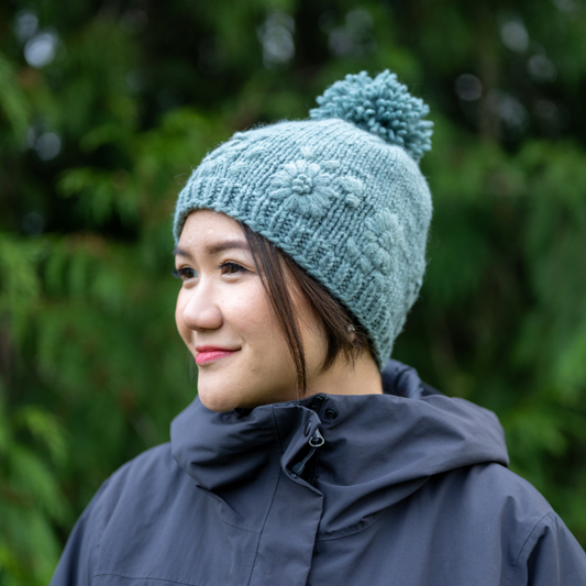 Woman wearing Ambler Petal handknit women's toque in eucalyptus with floral design and medium pom. 
