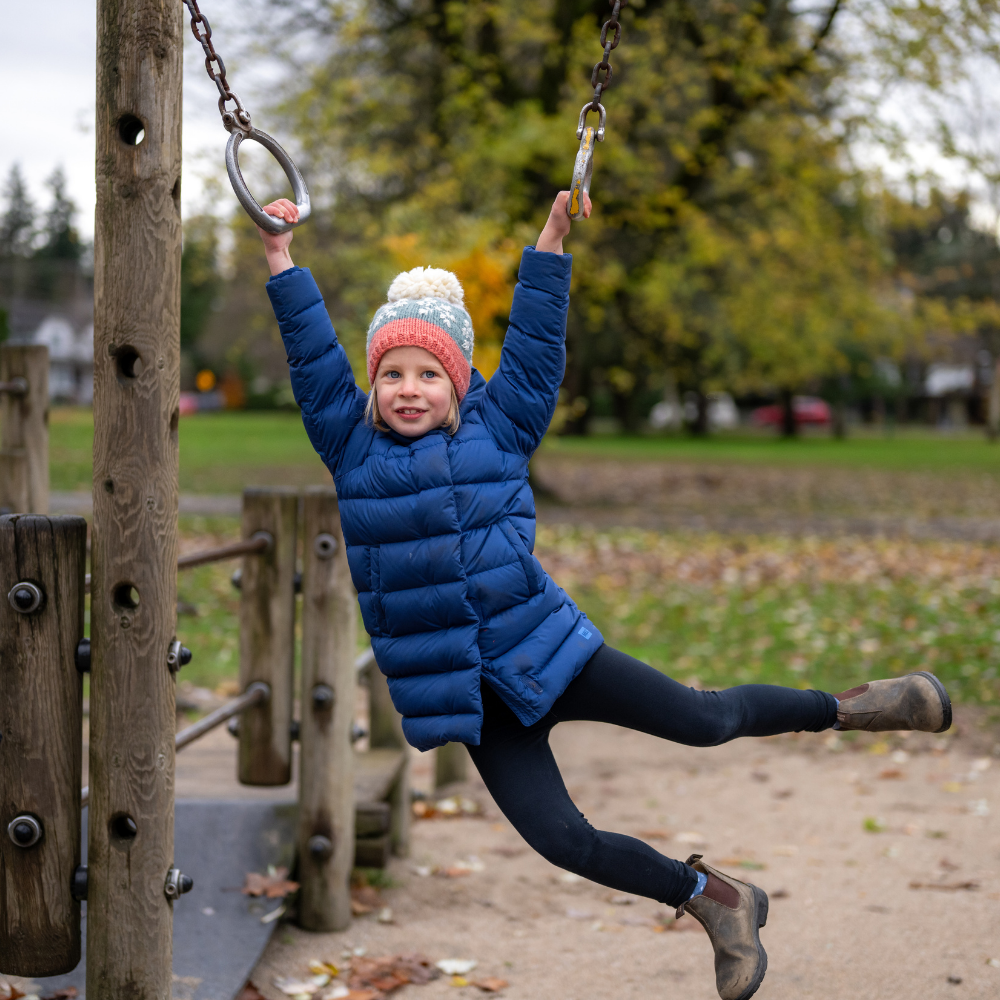 Girl wearing Ambler Snowflake handknit kids' toque in coral with embroidered snowflake design and medium pom. 