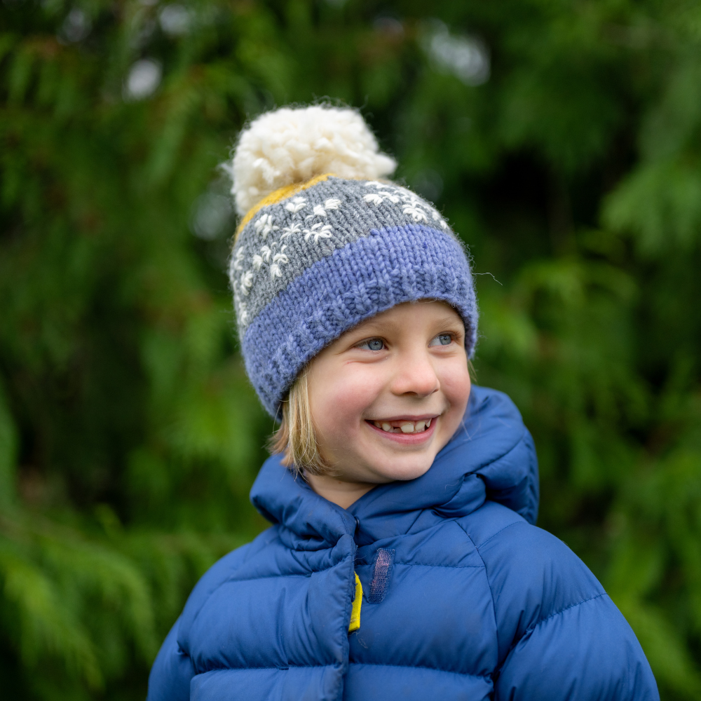 Girl wearing Ambler Snowflake handknit kids' toque in purple with embroidered snowflake design and medium pom. 