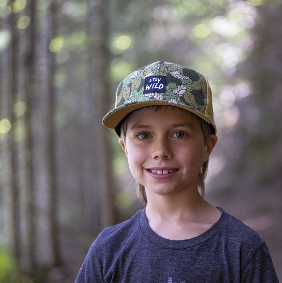 Boy wearing a kids&#39; hat with leaf pattern and &#39;Stay Wild&#39; patch on the front panel. 
