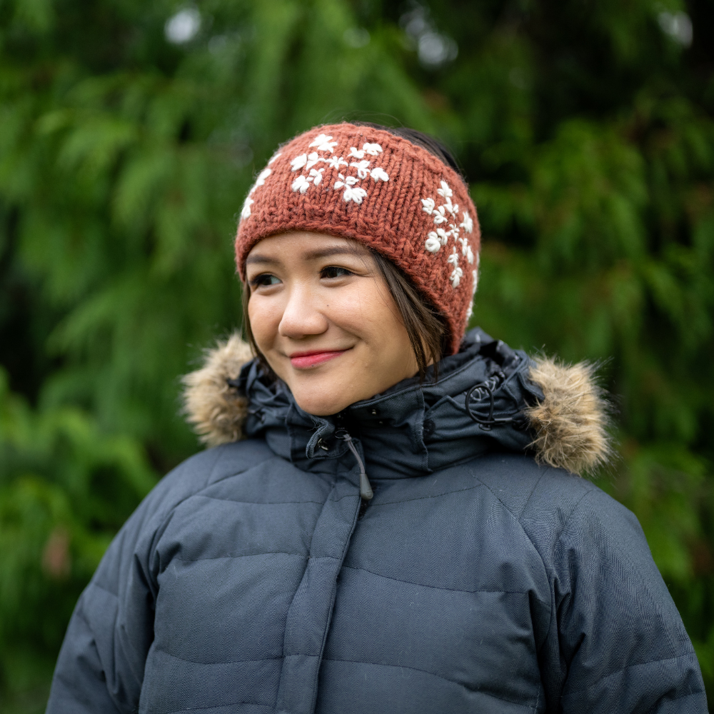 Woman wearing Ambler Stellar handknit winter headband in brick with snowflakes pattern and fleece lining. 