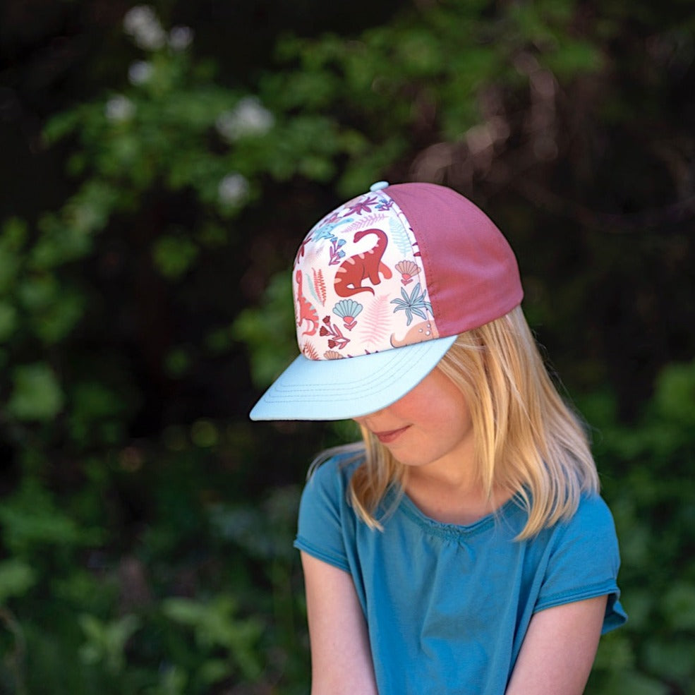 Girl wearing a pink children's cap with blue brim and colorful dinosaur pattern on the front panel. 