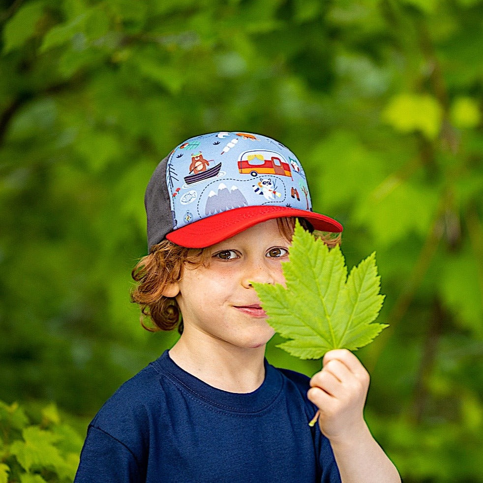 Boy wearing blue children's cap with red brim and colorful camping pattern on the front panel. 