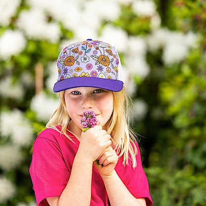 Girl wearing a purple kids' trucker hat with a dark purple brim, white mesh back and colourful floral pattern on the front panel.