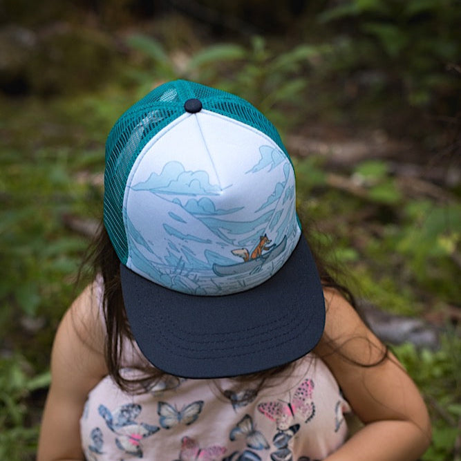 Toddler wearing trucker hat with teal back and fox in a canoe design on a white background. 